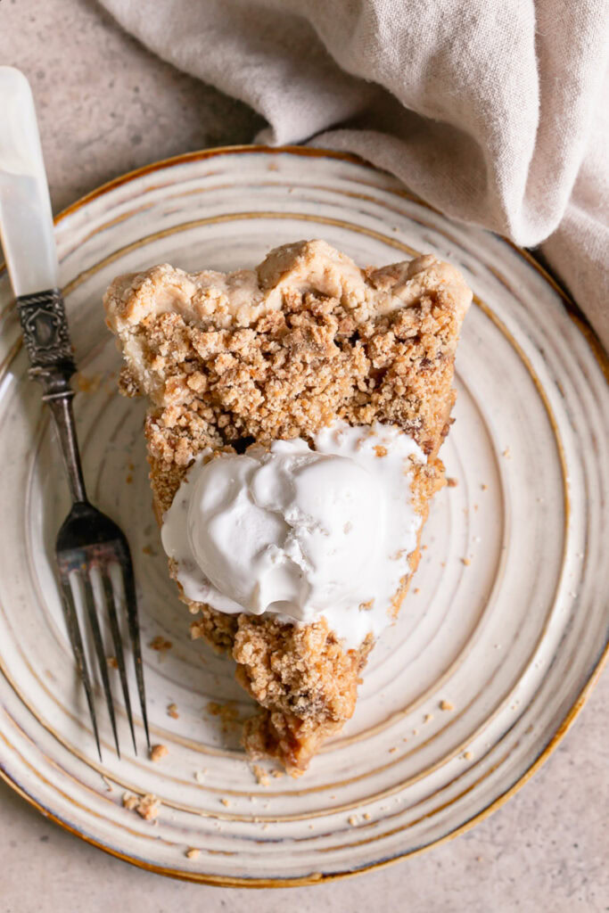 an overhead shot of a slice of apple pie on a plate with a fork and a scoop of vanilla ice cream on top