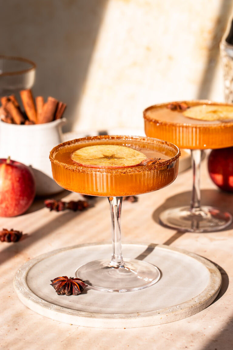 two glasses of pumpkin apple cider mocktail with a jar of cinnamon sticks, two apples, star anise in the background