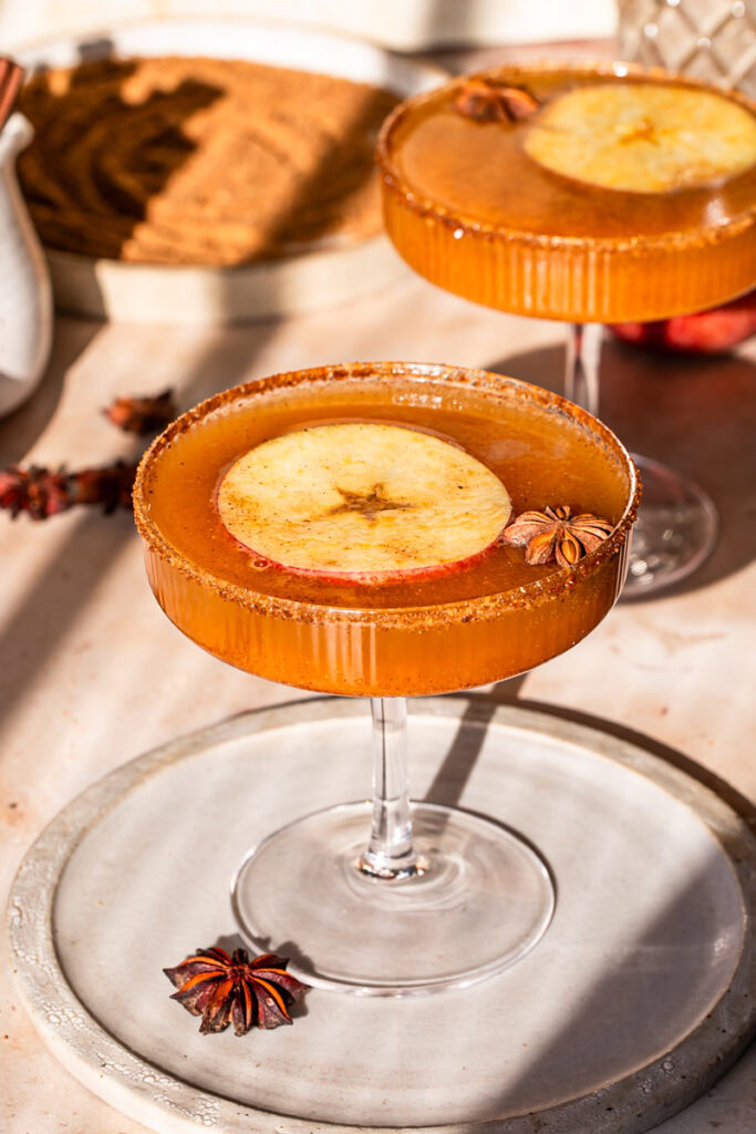 a close up of pumpkin apple cider mocktail with a slice of apple and star anise in the glass, a plate of coconut sugar in the background