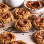 Pumpkin Spice Oatmeal Thumbprint Cookies with Date Caramel on a round cooling rack with a bowl of date caramel and a jug of oat milk and a cup of cinnamon sticks