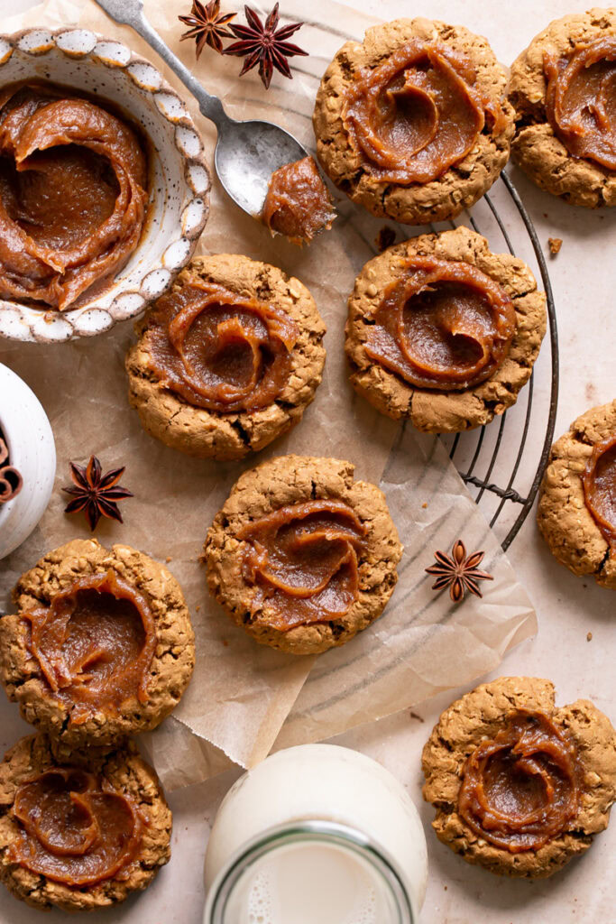 Pumpkin Spice Oatmeal Thumbprint Cookies with Date Caramel on a round cooling rack with a bowl of date caramel and a jug of oat milk