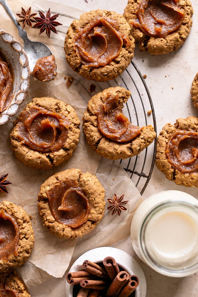 Pumpkin Spice Oatmeal Thumbprint Cookies with Date Caramel on a round cooling rack, one with a bite taken out of it and a bowl of date caramel and a jug of oat milk