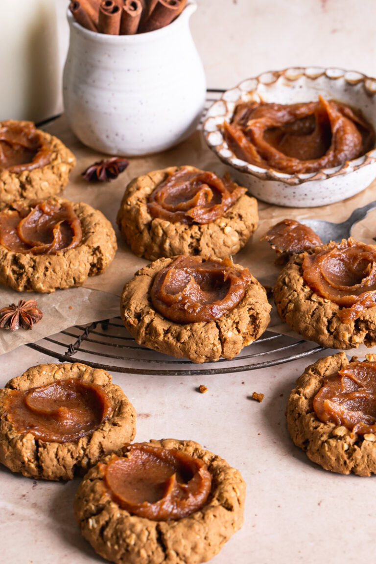 Pumpkin Spice Oatmeal Thumbprint Cookies with Date Caramel on a round cooling rack with a bowl of date caramel and a jug of oat milk and a cup of cinnamon sticks