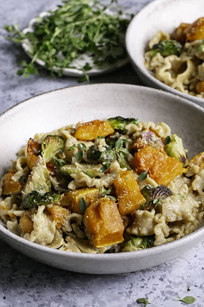 3/4 shot of two bowls of Buttery Butternut Squash and Brussels Sprouts Pasta on a gray blue stone backdrop with a small plate of fresh thyme in the background