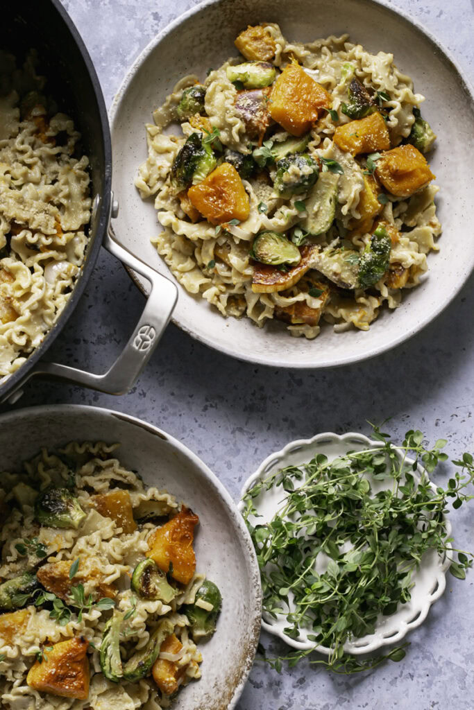 two bowls of Buttery Butternut Squash and Brussels Sprouts Pasta with a small plate of fresh thyme and the pot of pasta peeking in the frame from the left side