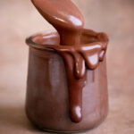 a jar of healthy vegan hot fudge, oozing over the side with a spoon scooping some hot fudge out.
