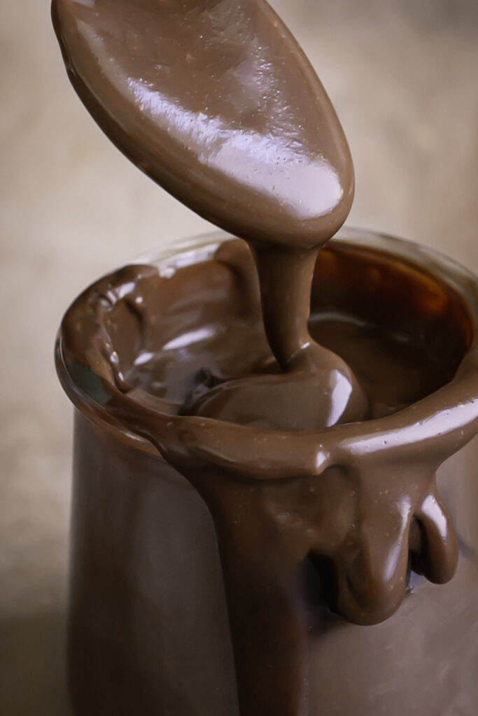 a close up of a jar of healthy vegan hot fudge, oozing over the side with a spoon scooping some hot fudge out.