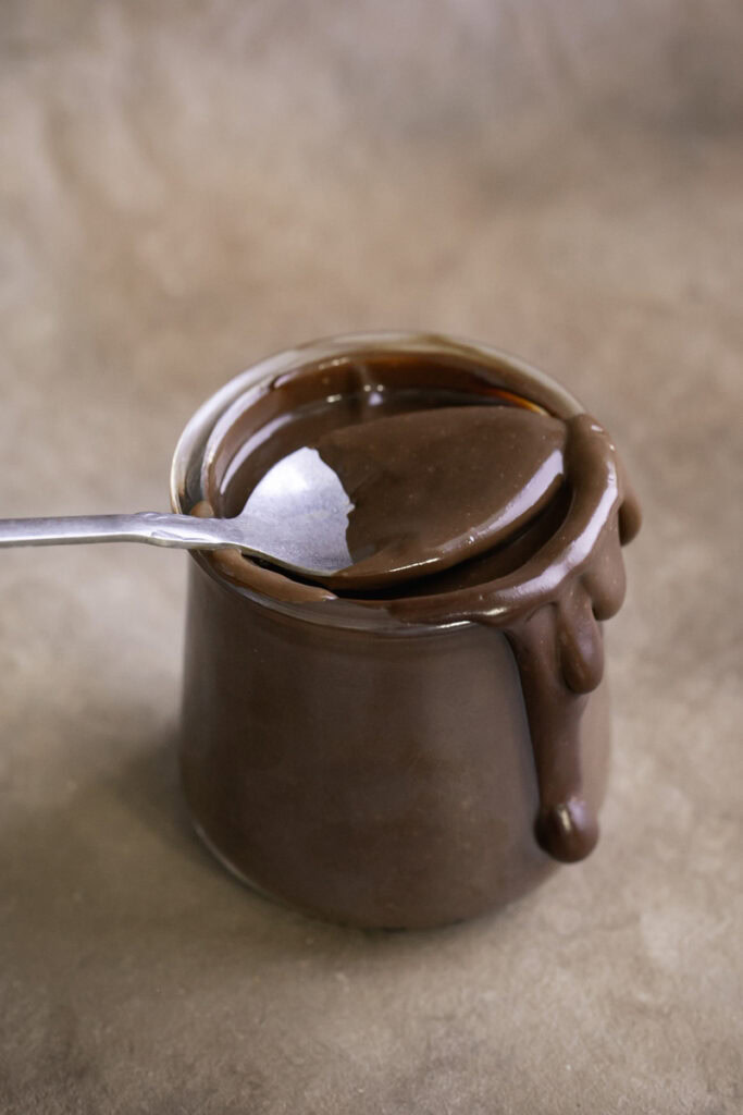 a jar of healthy vegan hot fudge, oozing over the side with a spoon resting on the mouth of the jar with hot fudge on it.