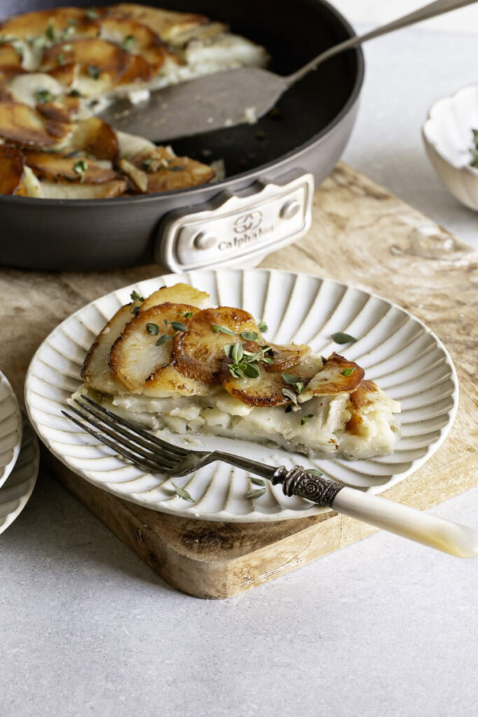 a slice of Pommes Anna skillet potatoes on a plate with a fork, the skillet of potatoes in the background