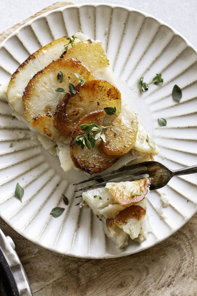 an overhead shot of a slice of Pommes Anna skillet potatoes garnished with fresh thyme, a fork slicing a piece of the potatoes