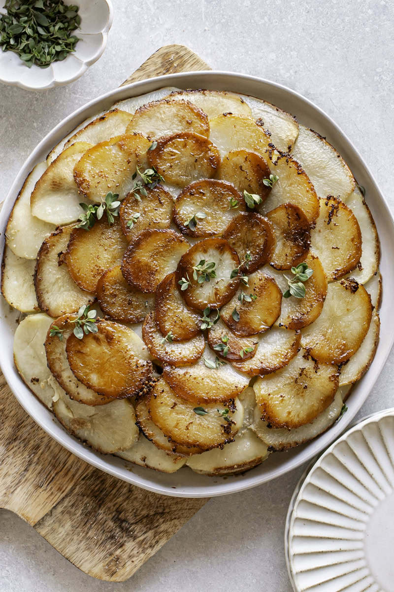 Pommes Anna skillet potatoes on a plate on top of a wooden cutting board, a bowl of fresh thyme in a small bowl above it