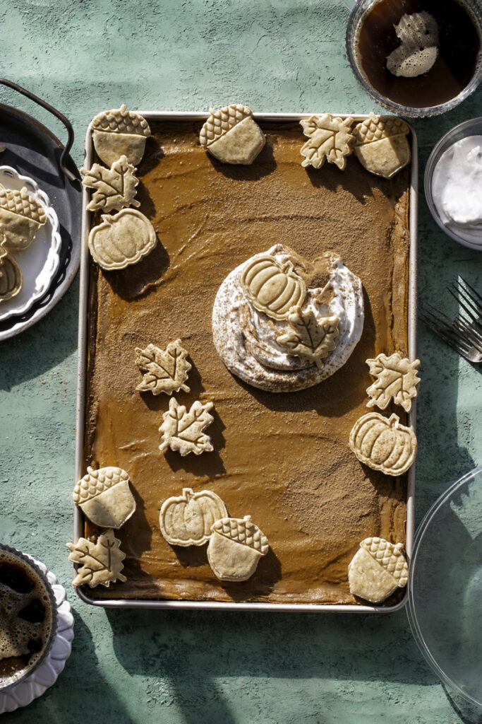 Sheet Pan Pumpkin Pie decorated with a dollop of whipped cream and pie crust cut outs. Two mugs of coffee, a bowl of whipped cream, and glass plates in the frame around the sheet pan pie