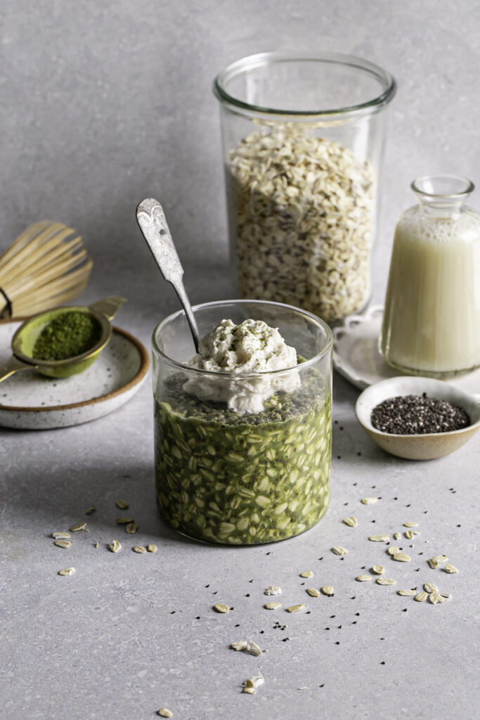 a glass of Oat Milk Matcha Latte Overnight Oats with a spoon sticking out of it topped with dairy-free whipped cream, a jar of oats, oatmilk, a bowl of chia seeds, a matcha whisk and a sifter with matcha in it in the background.