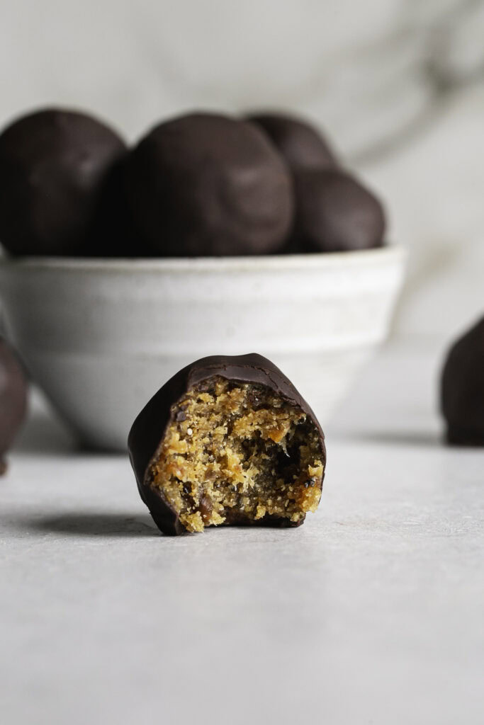 a single Chocolate covered Peanut Butter Almond Flour Date Bite with a bite taken out of it sitting in front of a bowl of Chocolate Peanut Butter Almond Flour Date Bites