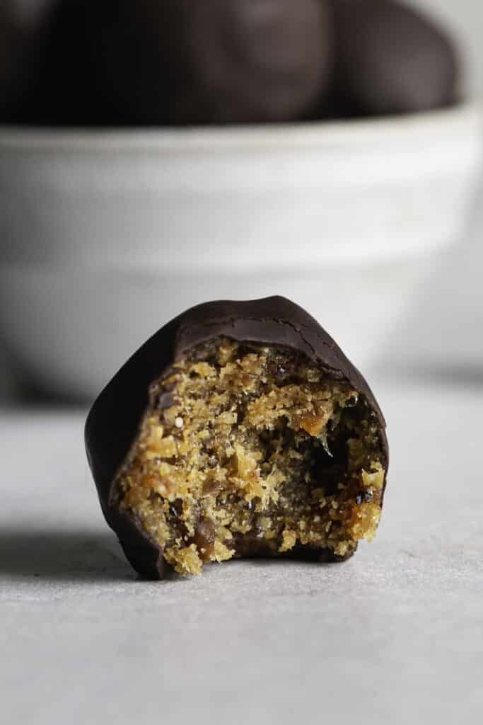 a close up of a single Chocolate covered Peanut Butter Almond Flour Date Bite with a bite taken out of it sitting in front of a bowl of Chocolate Peanut Butter Almond Flour Date Bites