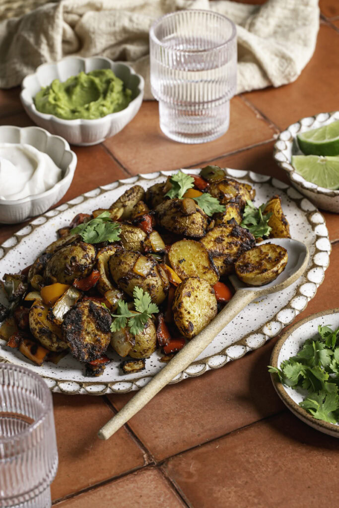 a side view of an oval ceramic plate with fajita roasted potatoes and a spoon. Small bowls of guacamole, vegan sour cream, lime slices, and fresh cilantro. A plate of soft corn tortillas, and two glasses of water.