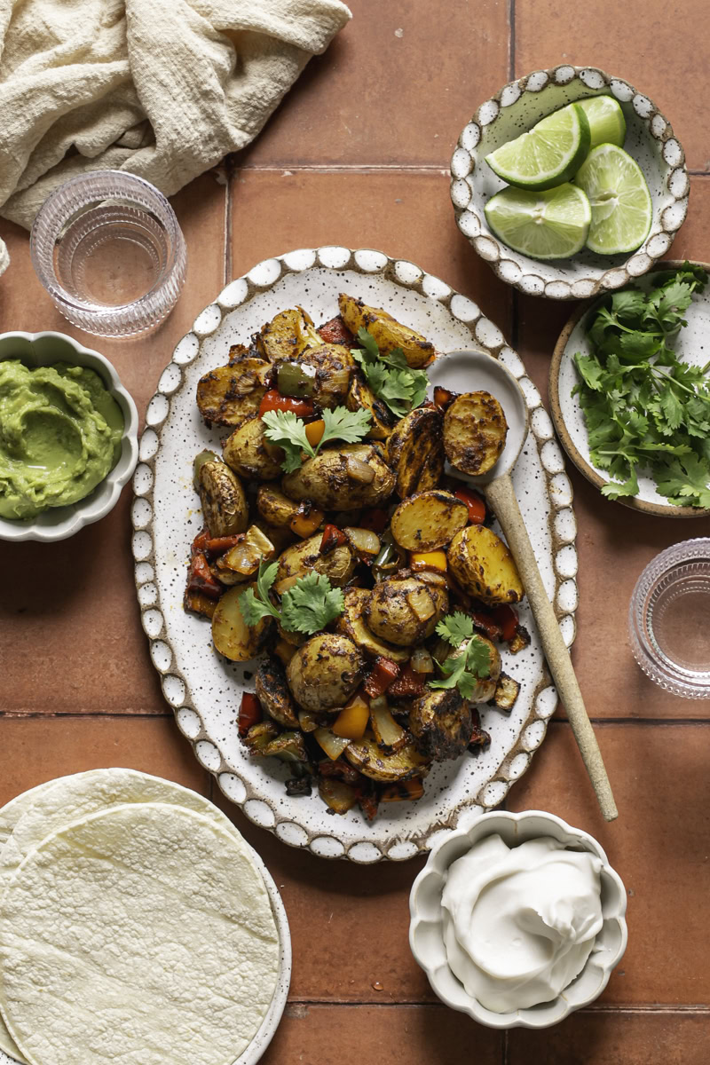 an oval ceramic plate with fajita roasted potatoes and a spoon. Small bowls of guacamole, vegan sour cream, lime slices, and fresh cilantro. A plate of soft corn tortillas, and two glasses of water.