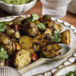 a side close up photo of fajita roasted potatoes on an oval ceramic plate with a spoon, a glass of water and a bowl of guacamole in the background.