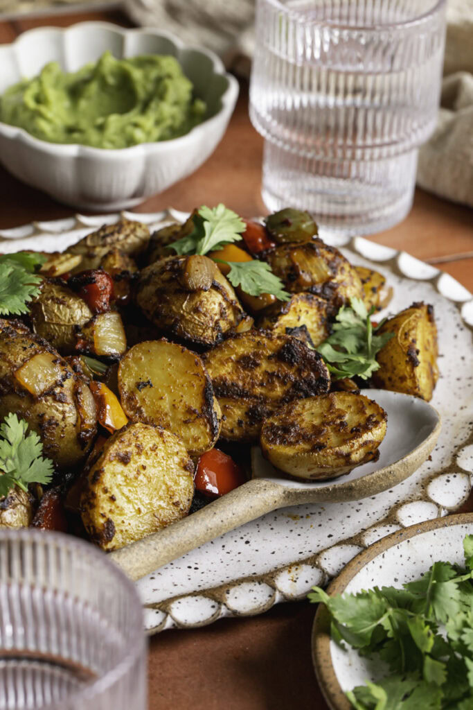 a side close up photo of fajita roasted potatoes on an oval ceramic plate with a spoon, a glass of water and a bowl of guacamole in the background.