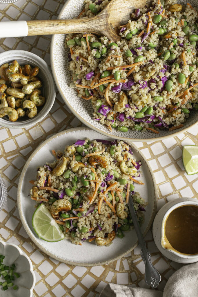 overhead shot of Edamame Quinoa Salad with a fresh lime slice on the plate, a bowl of salad with a spoon in it in the background, a jar of peanut ginger Lime Dressing, a bowl of sliced green onion, and a bowl of agave lime roasted cashews.