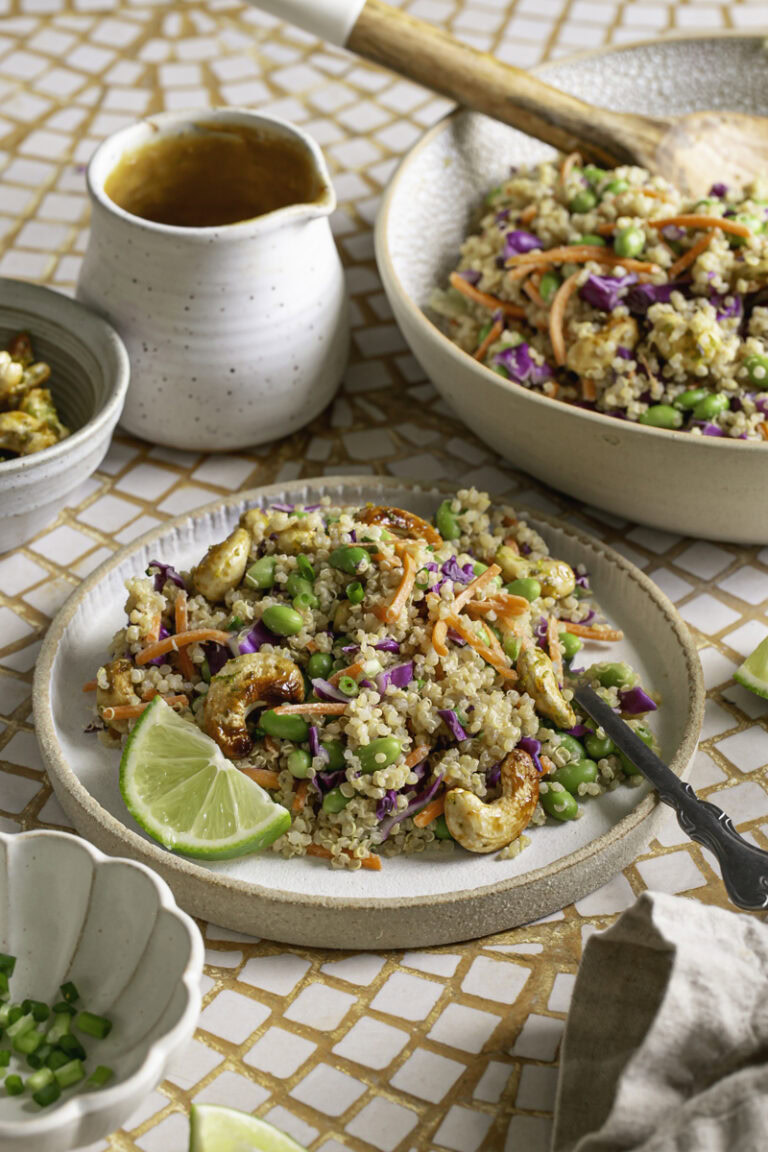 a plate of Edamame Quinoa Salad with a fresh lime slice on the plate, a bowl of salad with a spoon in it in the background, a jar of peanut ginger Lime Dressing, a bowl of sliced green onion, and a bowl of agave lime roasted cashews.