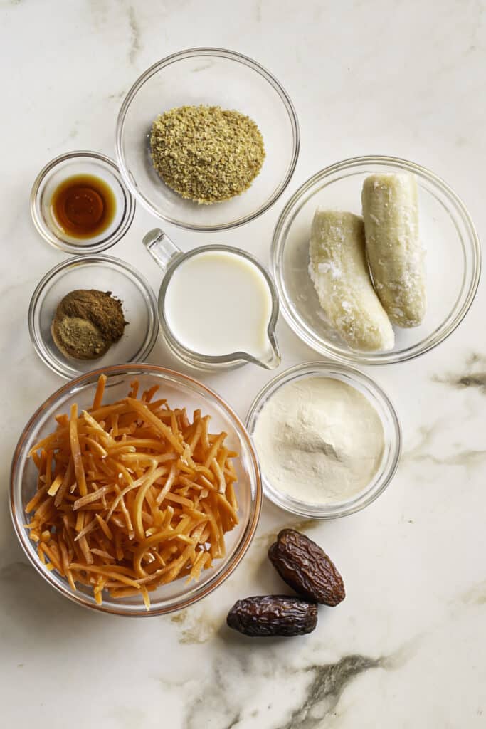 all of the ingredients separated into bowls that are used to make this carrot cake protein smoothie: shredded carrots, frozen banana, flaxseed meal, oat milk, dates, plant protein powder, vanilla, and spices.