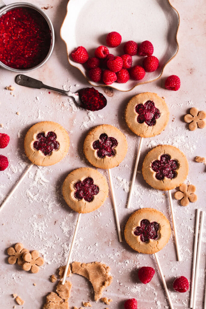 six raspberry pie pops laid out with a plate of fresh raspberries and a pot of raspberry jam beside it