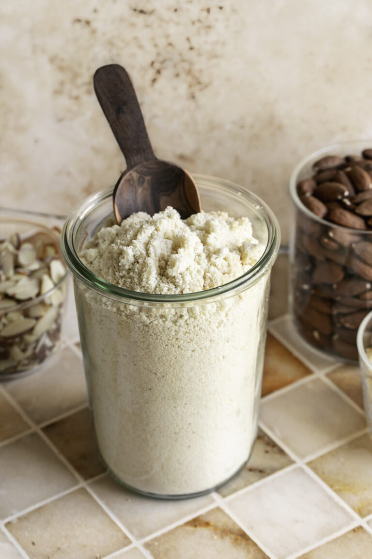 almond flour in a glass jar with a small wooden scoop in it and whole and sliced almonds in glasses behind it.