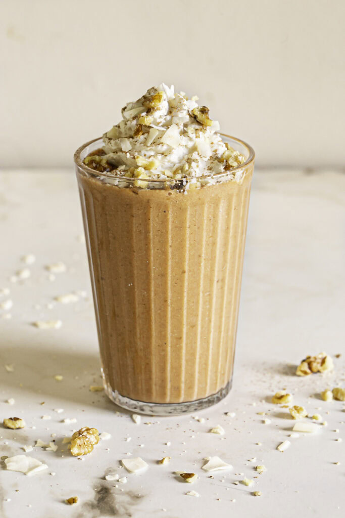 a glass of carrot cake protein smoothie topped with whipped cream, chopped walnuts, and toasted coconut chips.
