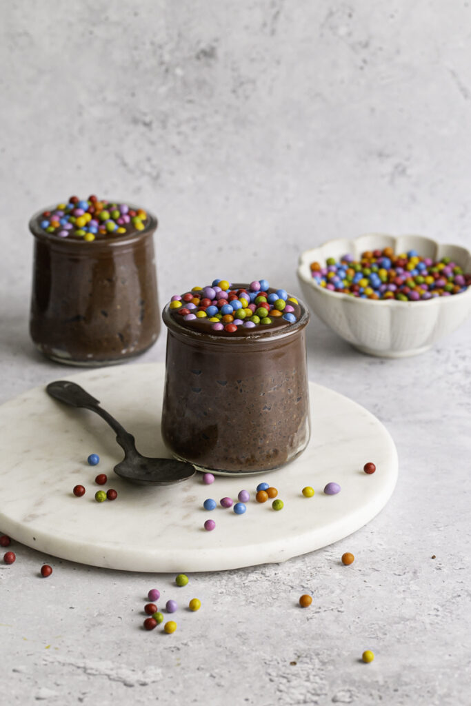 two jars of Cosmic Brownie Chocolate Chia Seed Pudding with a bowl of candy coated rainbow chocolate chips in the background.