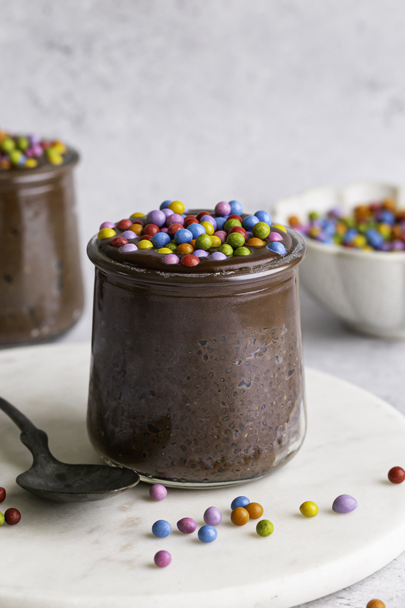 close up shot of a jar of Cosmic Brownie Chocolate Chia Seed Pudding with a bowl of candy coated rainbow chocolate chips and another jar of chia pudding in the background.