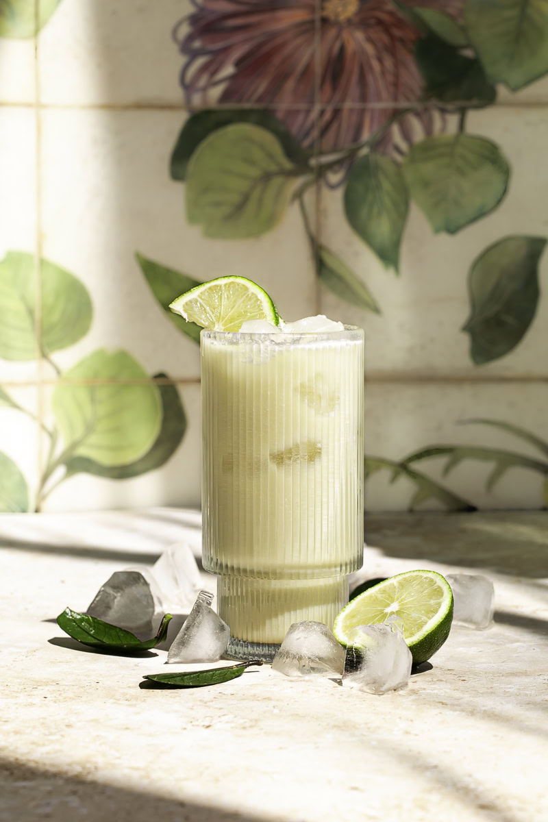 a glass of creamy coconut milk brazilian lemonade garnished with lime slices, half a cut lime at the base of the glass and ice cubes surrounding the glass.