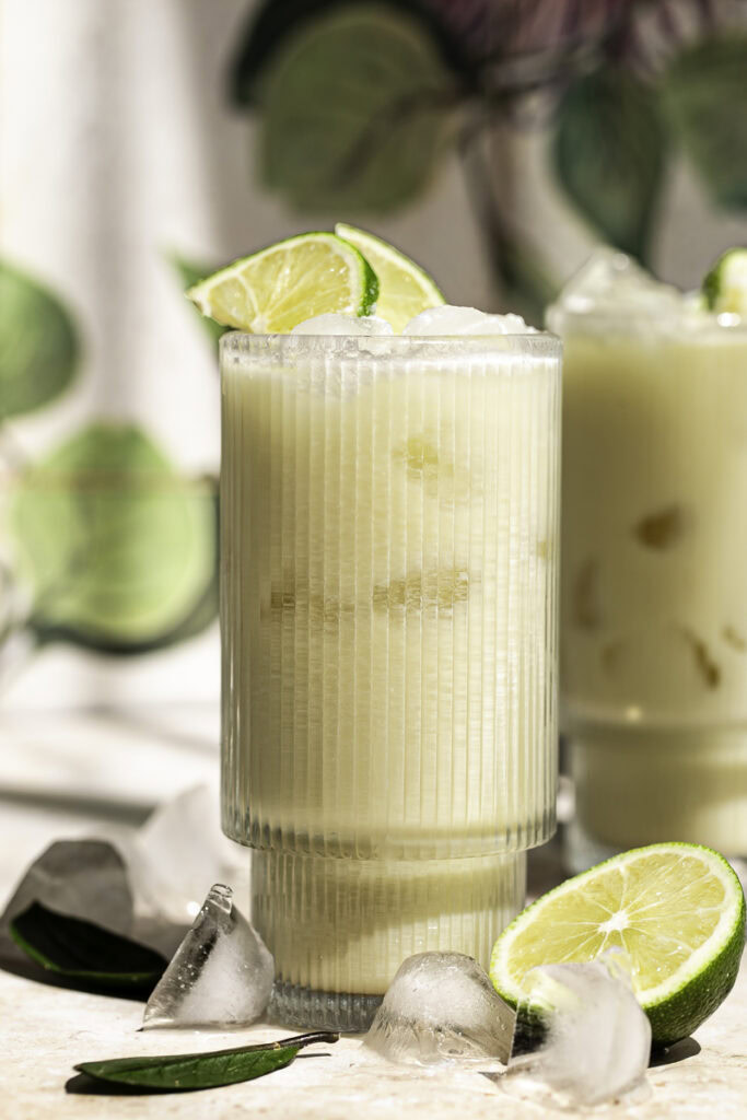 a close up shot of two a glasses of creamy coconut milk brazilian lemonade garnished with lime slices, half a cut lime at the base of the glass and ice cubes surrounding the glass.