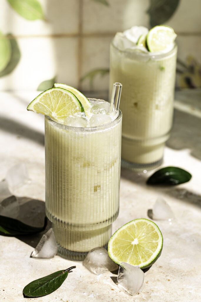 3/4 shot of two glasses of creamy coconut milk brazilian lemonade garnished with lime slices, half a cut lime at the base of the glass and ice cubes surrounding the glass.