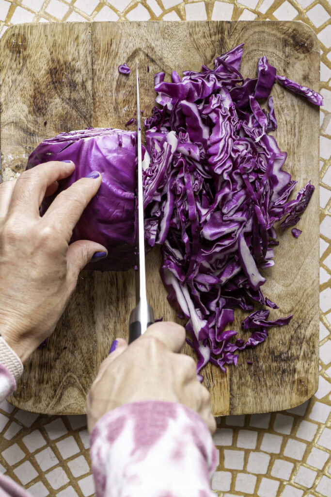 hand thinly slicing red cabbage into shreds on a wooden cutting board.