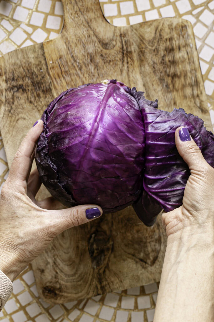 action shot of hands removing the outer leaf of a red cabbage that's sitting on top of a wooden cutting board.
