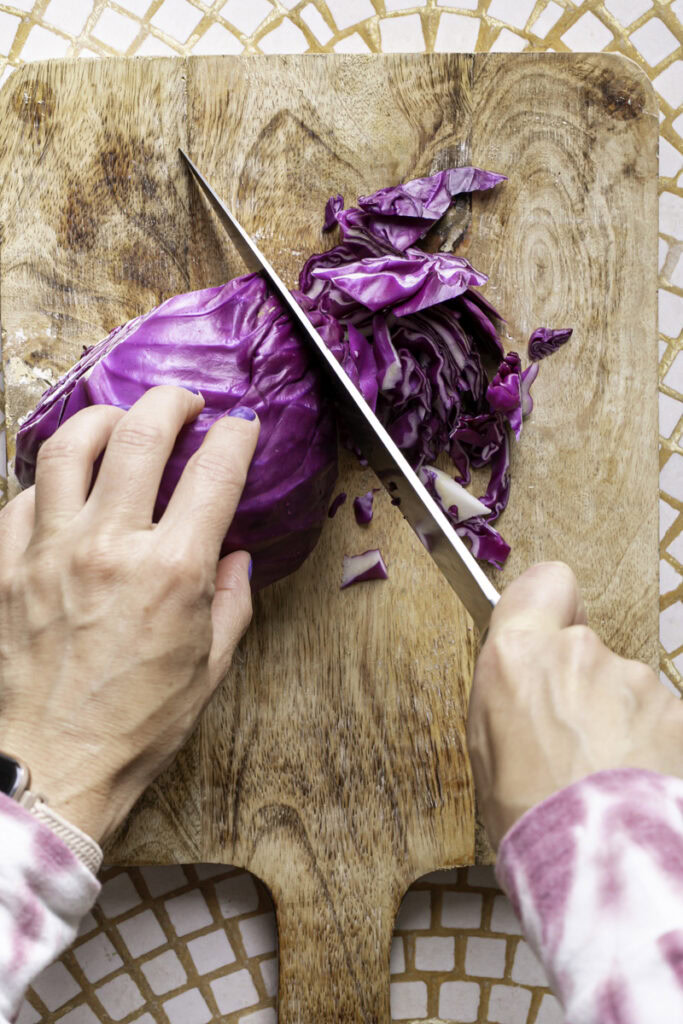 hands cutting a half of a head of cabbage with a large knife on a wooden cutting board, rotating the cabbage clockwise.