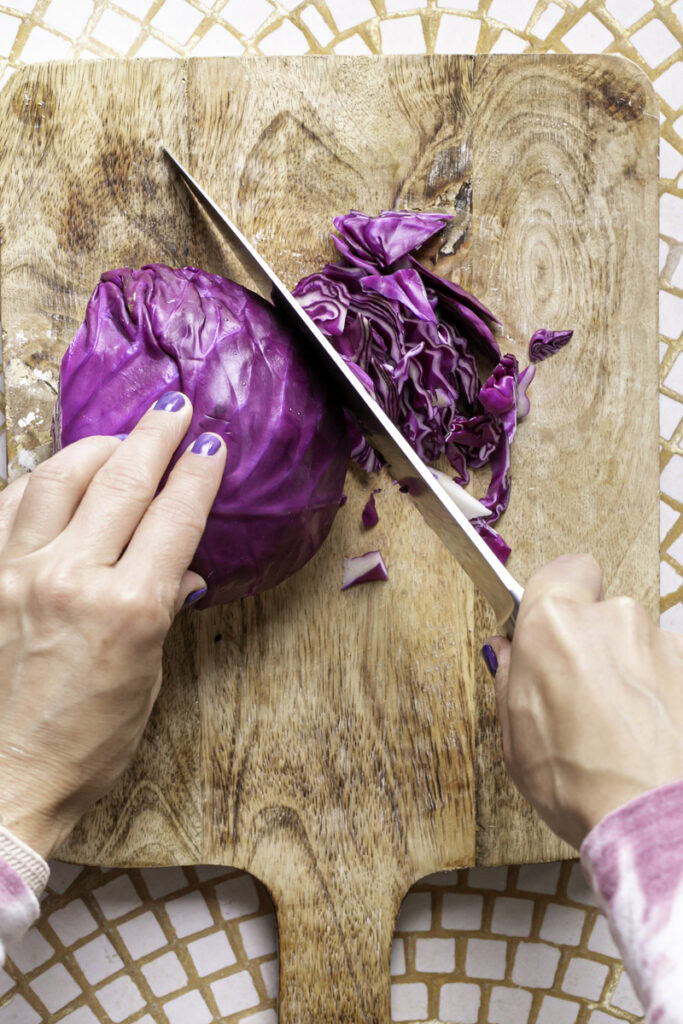 hands cutting a half of a head of cabbage with a large knife on a wooden cutting board.