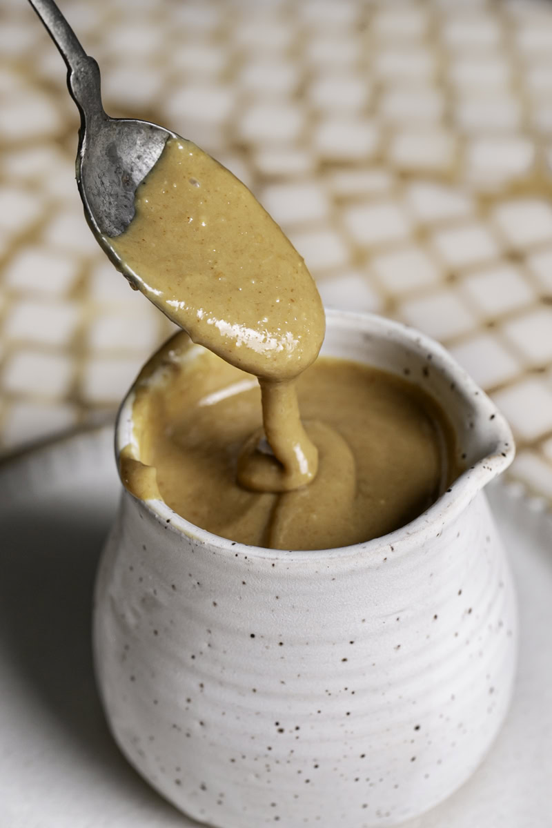 a close up of peanut ginger lime dressing in a ceramic dressing carafe with a spoon drizzling some of the dressing back into the carafe.