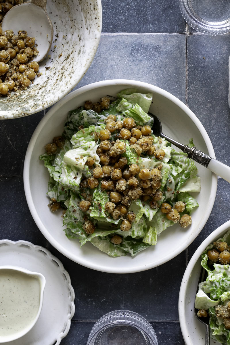 a bowl of romaine caesar salad topped with crispy chickpea croutons, another salad bowl in the bottom right corner, extra caesar dressing in the bottom left corner, and more crispy chickpeas in a bowl in the upper left corner.