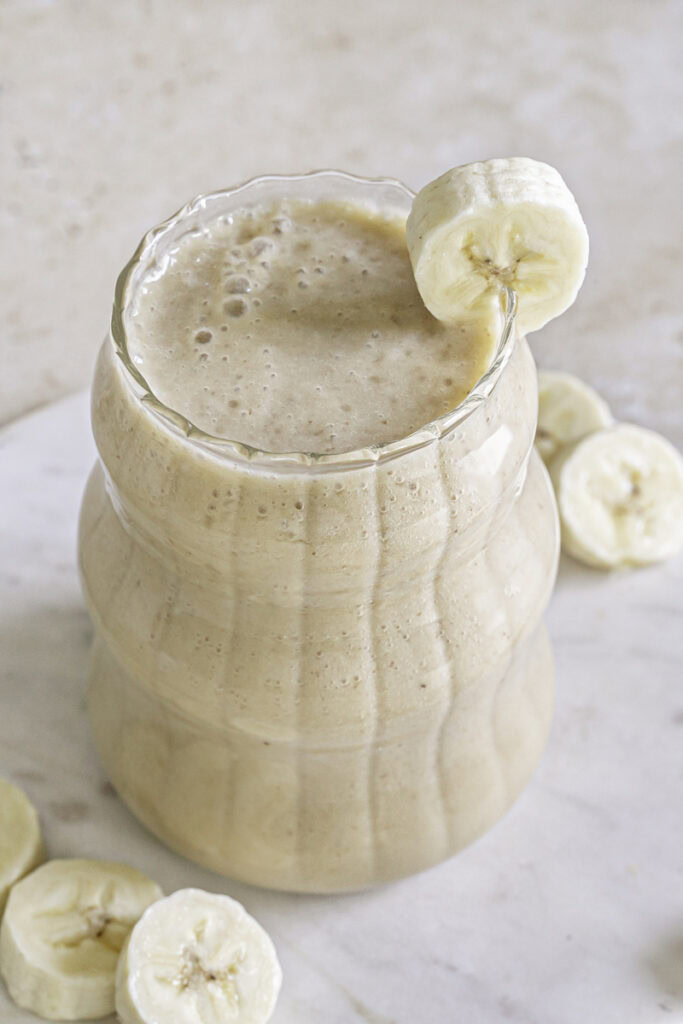 3/4 photo of banana milk in a ribbed bubble glass garnished with a slice of banana on the rim of the glass.