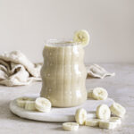 banana milk in a glass on top of a round marble plate with banana slices around it and a banana slice on the rim of the glass.