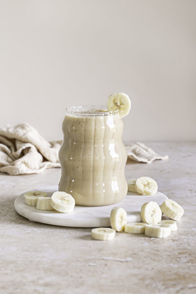 banana milk in a glass on top of a round marble plate with banana slices around it and a banana slice on the rim of the glass.
