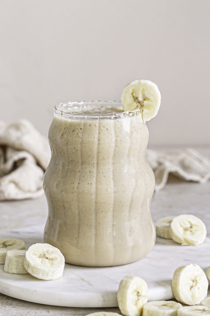 a close up image of banana milk in a glass on top of a round marble plate with banana slices around it and a banana slice on the rim of the glass.