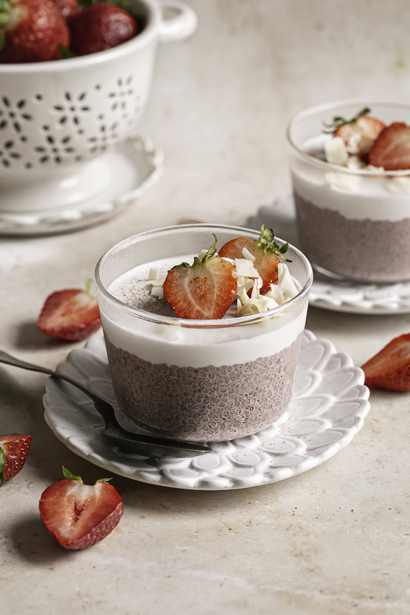 a glass filled with strawberry hibiscus chia seed pudding topped with yogurt, fresh strawberries and toasted coconut chips with another chia pudding in the background.