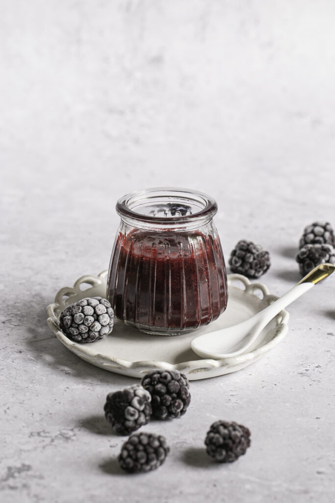 a small glass jar of blackberry curd with frozen blackberries around it and a small spoon resting on a plate next to the jar.