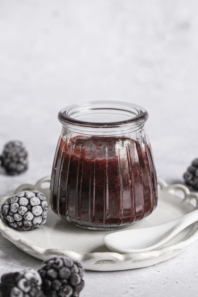 a close up image of a small glass jar of blackberry curd with frozen blackberries around it and a small spoon resting on a plate next to the jar.