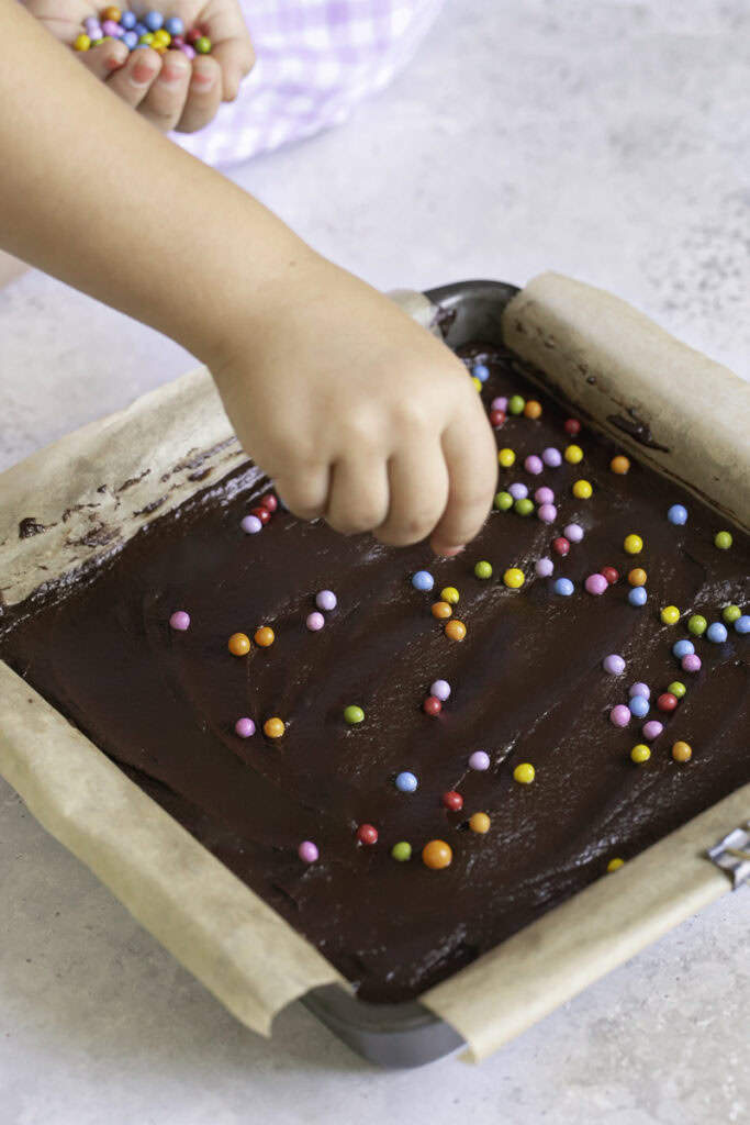 action shot sprinkling the rainbow candy coated chocolate chips on top of the chocolate ganache frosting.