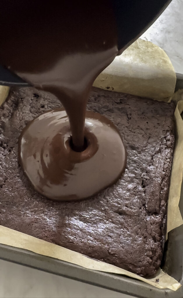 action shot pouring melted chocolate ganache on top of the baked brownies.