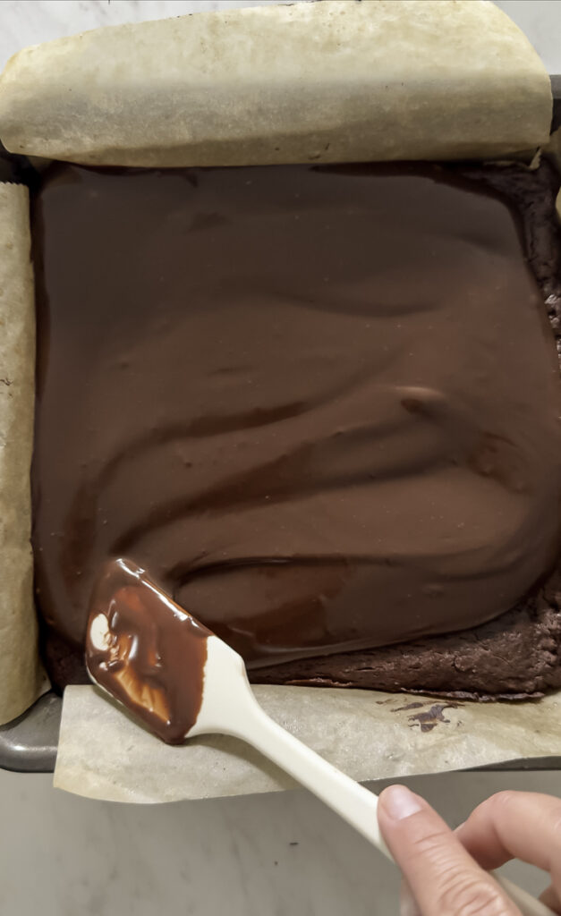 action shot spreading the chocolate ganache over the brownies using a spatula.